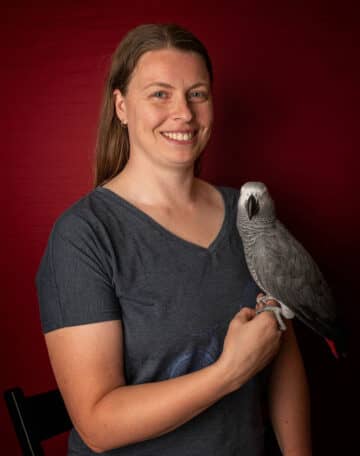 A grey parrot sits on a woman's hand, the two are looking at the camera